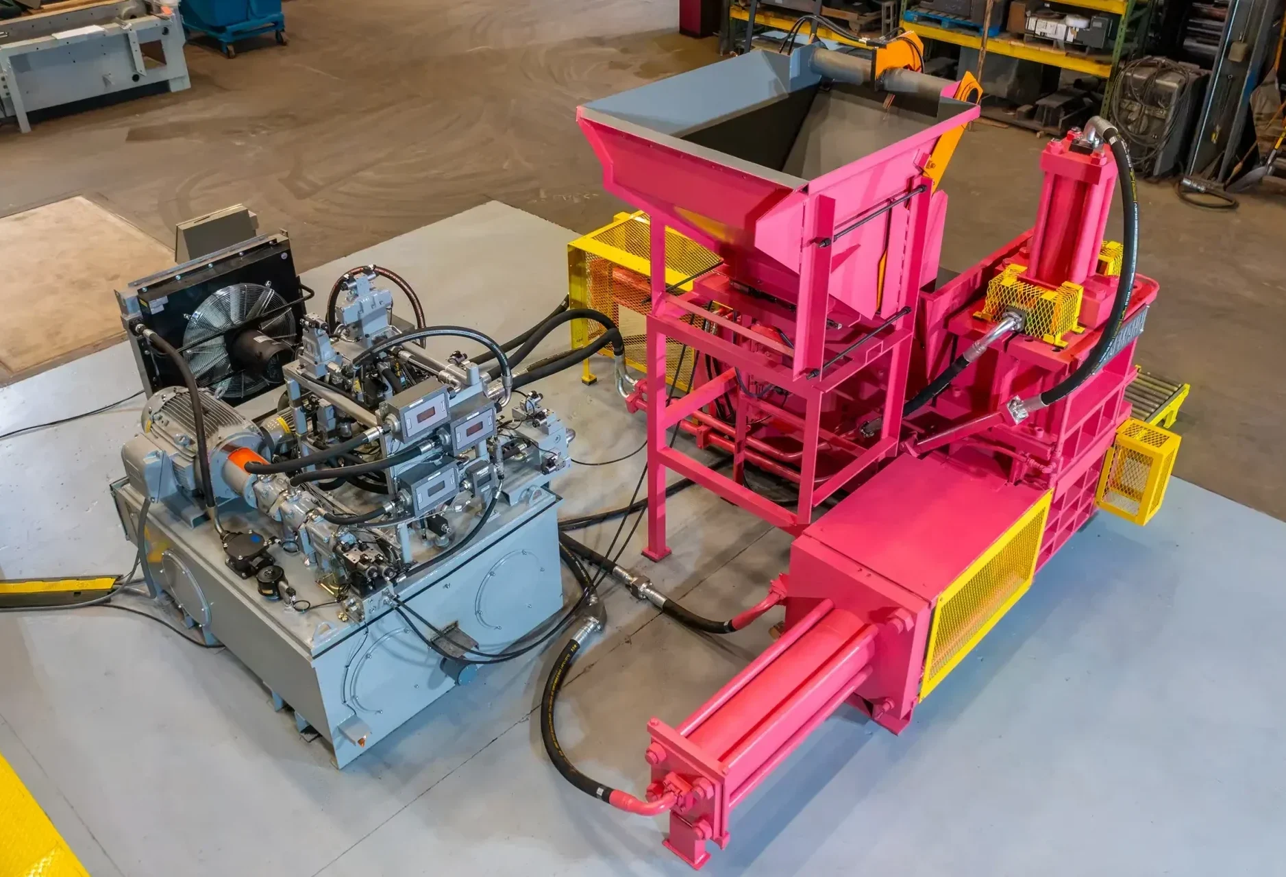 A large piece of industrial scrap baling equipment with pink and yellow components is connected to a gray hydraulic power unit by black hoses, set in a spacious workshop with various equipment and storage shelves in the background.