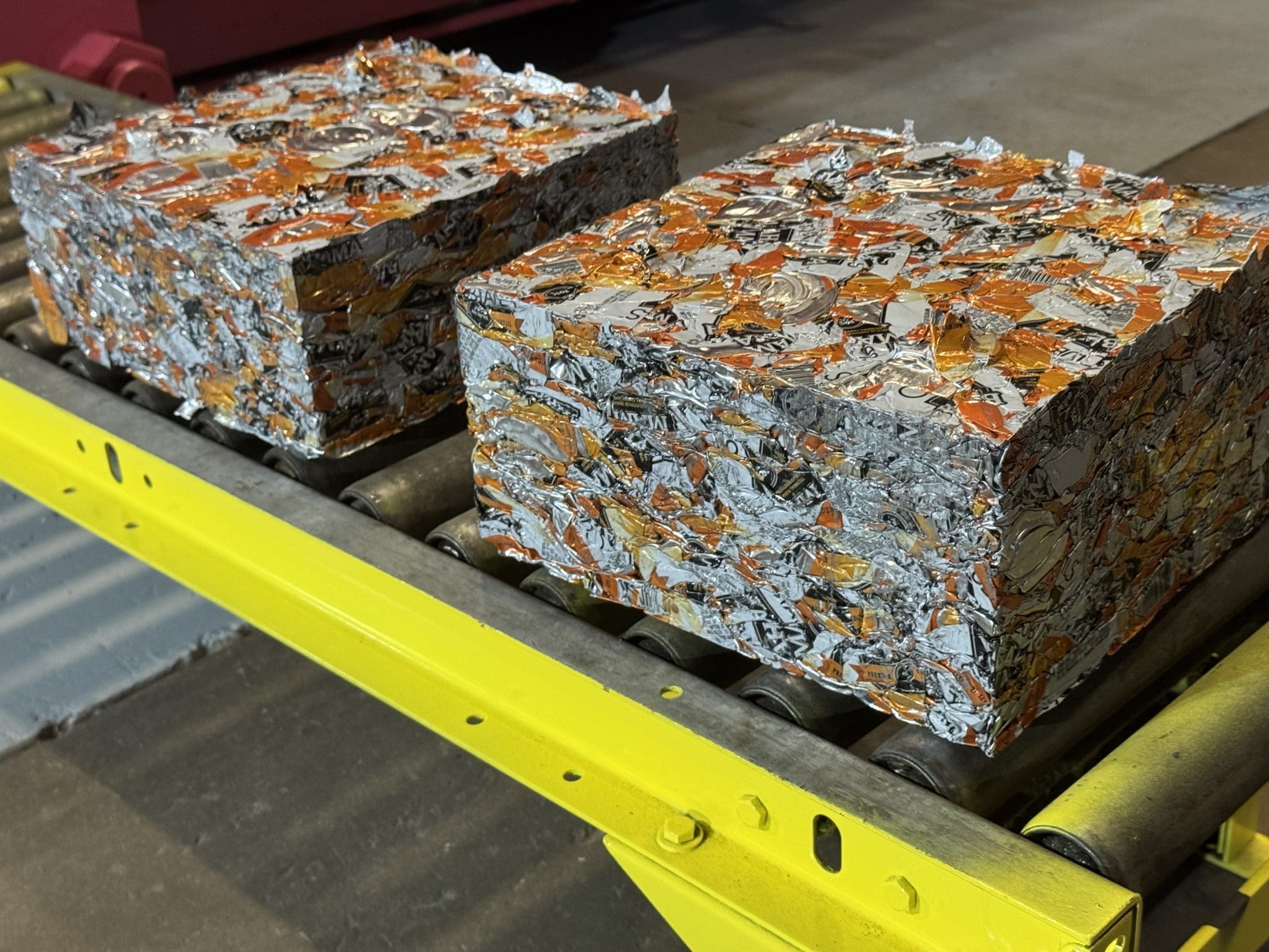 Two tightly compressed cubes of shredded aluminum cans, primarily silver and orange, demonstrate typical scrap material behavior as they sit on a yellow metal conveyor belt inside an industrial facility.