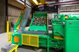 A green and yellow 2-ram baler in a factory processes and compresses scrap metal cubes, with a conveyor feeding the metal into the machine amid a clean, organized workspace.