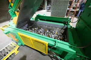 A large green and yellow 2-ram baler filled with numerous empty metal cans, located in a factory setting with shelves and other equipment visible in the background.