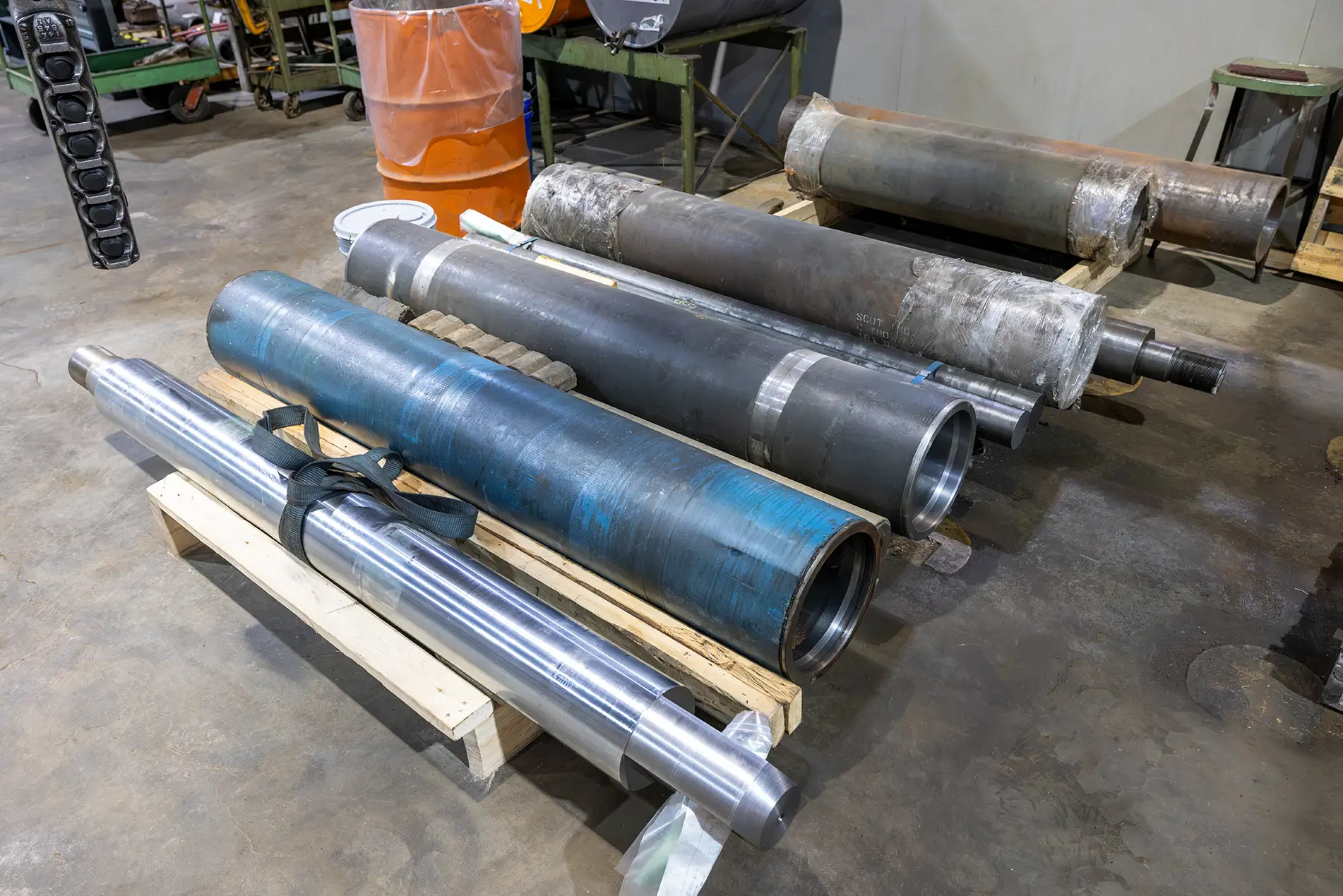 Large metal industrial rollers and cylinders—often used as industrial baler parts—are placed on wooden pallets in a workshop with a concrete floor, surrounded by equipment and tools, ready for maintenance or service.