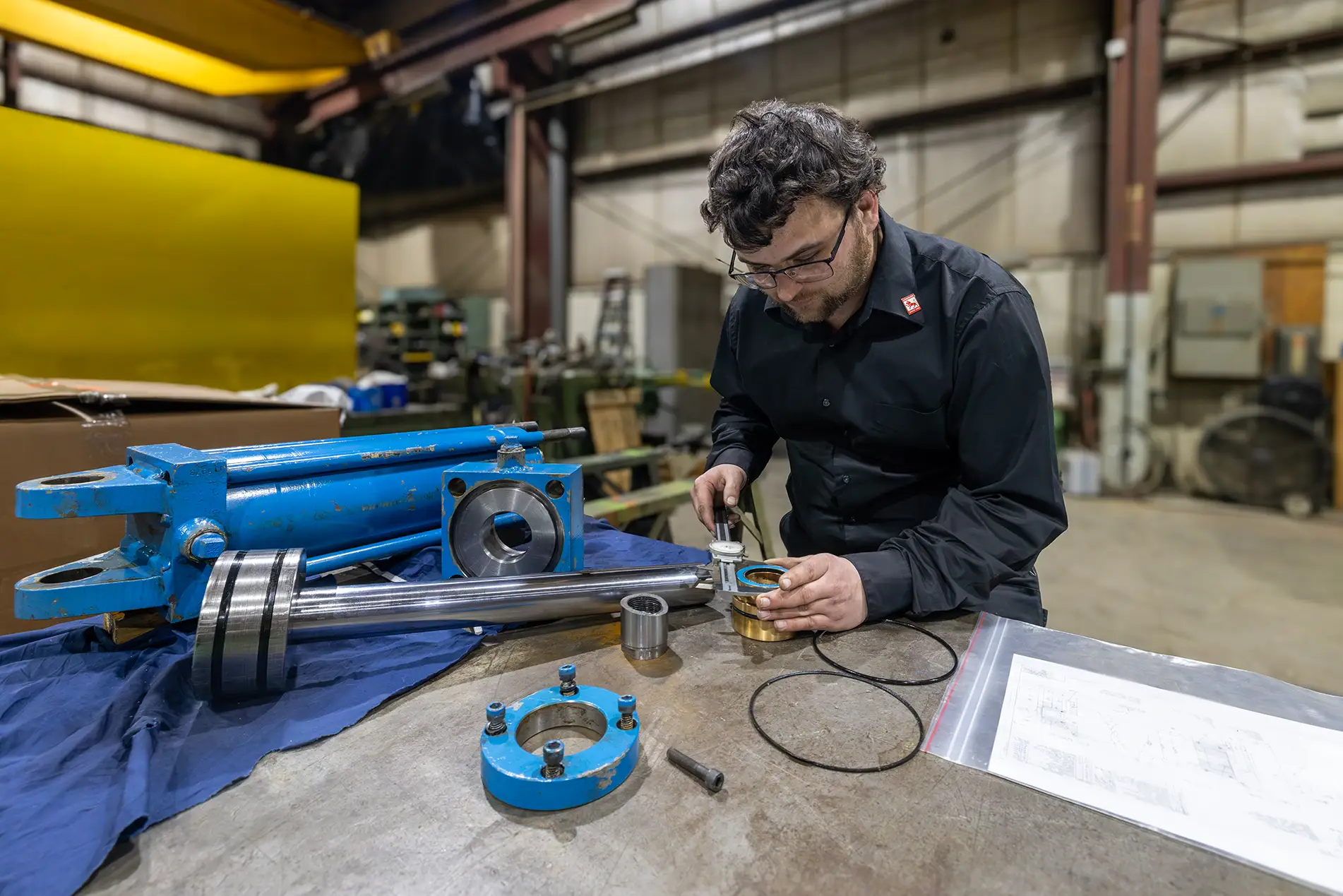 A man in a black shirt inspects metal mechanical parts with a caliper on a workbench in an industrial workshop, surrounded by large blue machine components, technical drawings, and industrial baler parts service tools.