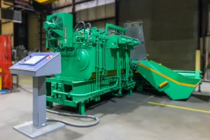 A large green 2-ram baler with various pipes and components stands in a factory. An angled chute extends from the side, and a separate control panel with buttons and a screen is positioned in the foreground.