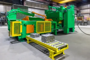 A green and yellow 2-ram baler compresses scrap metal into compact rectangular bales, which are placed on a conveyor in a spacious factory environment.