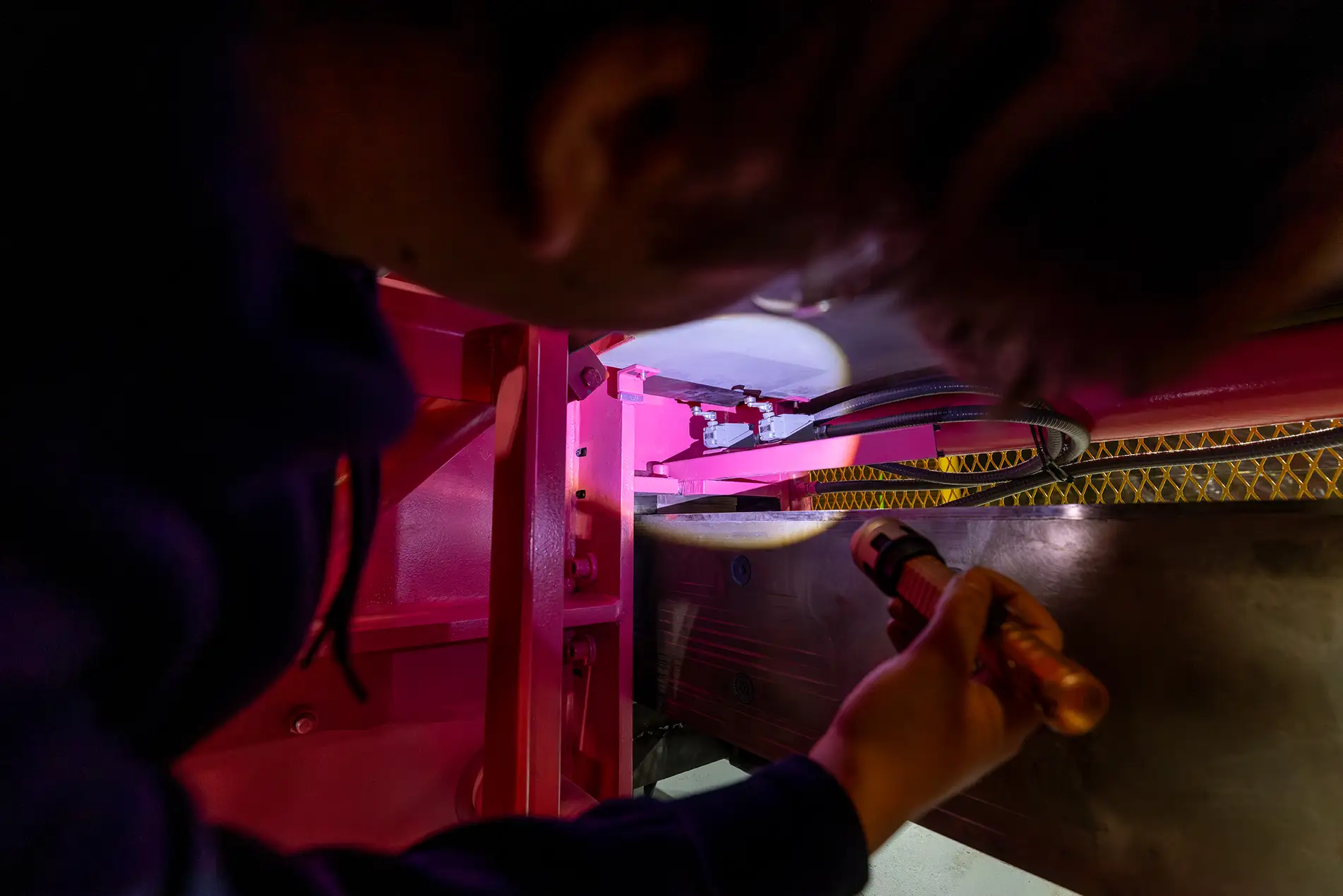 A person uses a flashlight to inspect machinery under pink lighting, focusing closely on wires and metal components during an industrial baler parts service in a mechanical setting.