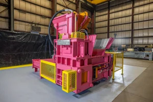 A large piece of industrial scrap baling equipment painted pink and yellow stands in a spacious warehouse with high ceilings and metal walls. The machine features various hoses, guards, and hydraulic components.