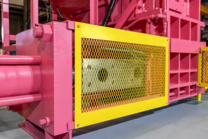 Close-up of a pink industrial scrap baling equipment machine with a yellow metal mesh safety guard covering its moving parts. The background features additional machine components, and the setting appears to be indoors.