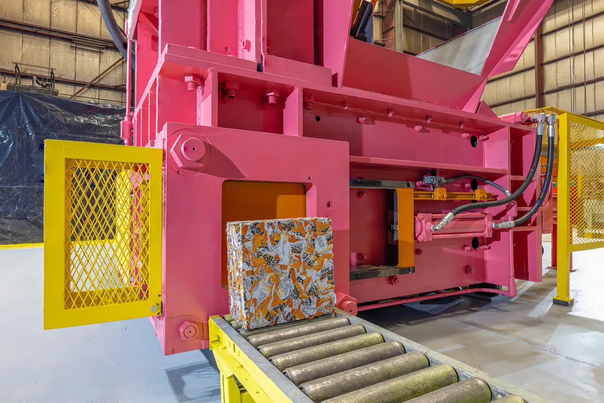 A compacted bale of scrap metal, shaped by the baling process and scrap material behavior, sits on rollers before a large pink industrial baler inside a factory with yellow safety barriers and metal walls.