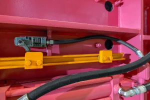 A close-up of a 3-ram metal baler painted pink and yellow, featuring a black sensor with wiring, a yellow metal rail, and black hoses connected to the equipment.