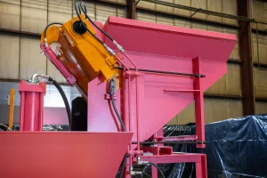 A close-up view of industrial metal balers with pink and orange components inside a factory, highlighting hydraulic systems and angled surfaces.