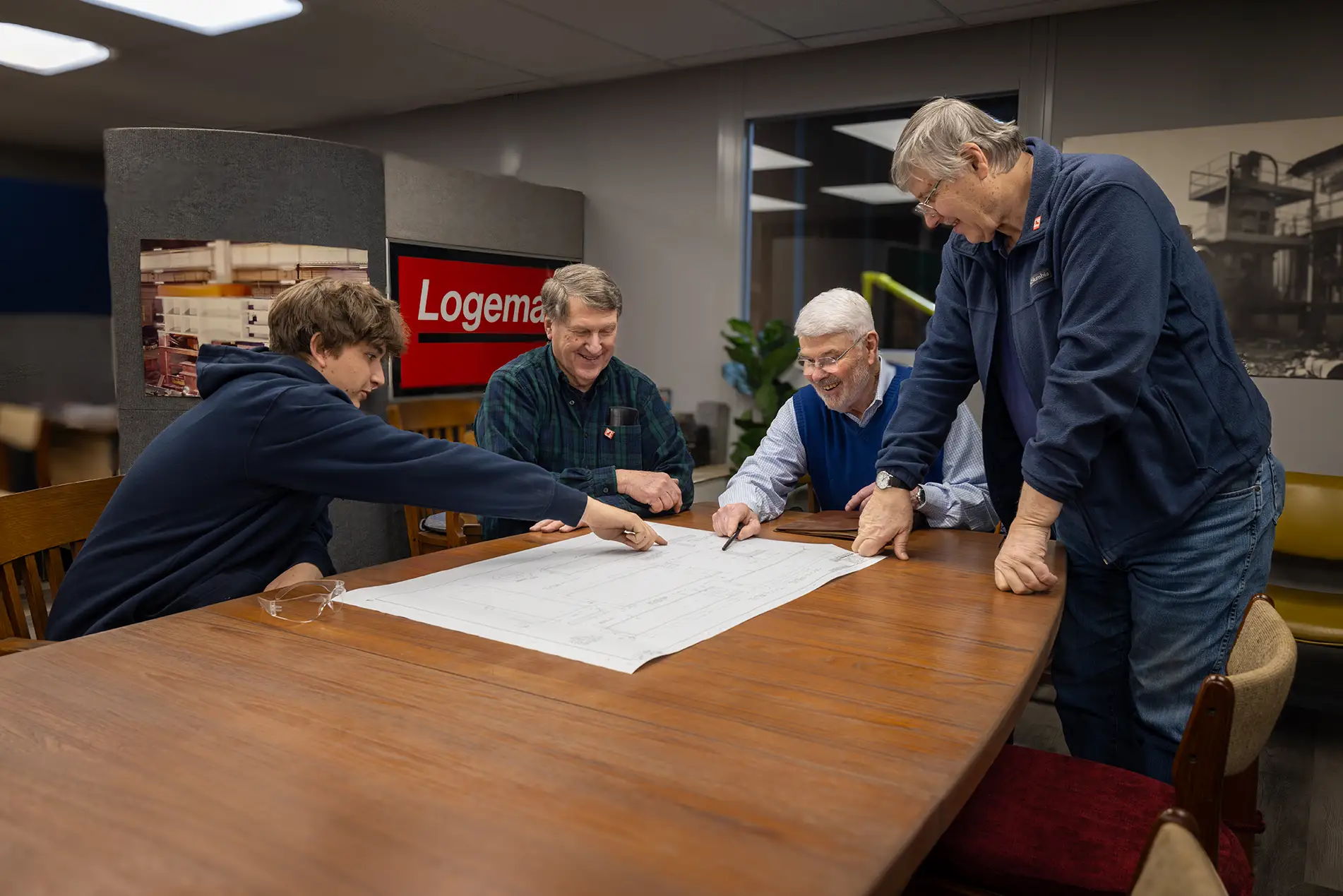 Four men gather around a wooden table, examining a large blueprint. They appear engaged and collaborative in a well-lit office space, with a Logemann sign in the background—ideal for those seeking metal baler manufacturer contact details.
