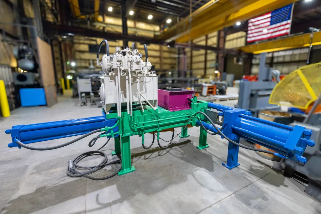 A brightly colored industrial hydraulic machine, resembling refurbished metal balers with blue and green components, hoses, and control units, stands inside a large factory with an American flag on the wall in the background.