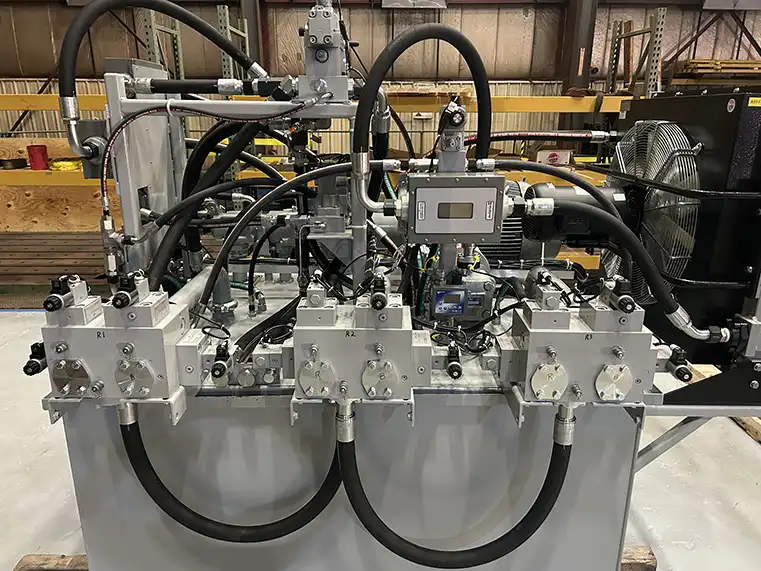 A hydraulic power unit baler with multiple control valves, hoses, and gauges is set up in an industrial warehouse. The unit features labeled ports, electronic controls, and connected piping. Shelves and equipment are visible in the background.